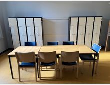 Ein einfacher Raum mit gelben Bodenfliesen, einem Holztisch und sechs Holzstühlen mit blauen Sitzpolstern. Im Hintergrund steht eine Reihe weißer Spinde mit schwarzen Rahmen an der Wand.