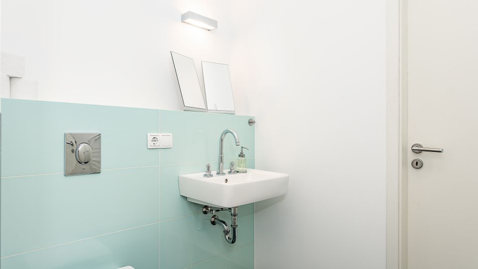 Kleines, helles Badezimmer mit wandmontiertem Waschbecken, verchromter Armatur und Mintgrün-Fliesen als Wandverkleidung. Über dem Becken hängt eine rechteckige Wandleuchte, daneben ist eine weiße Tür zu sehen.