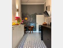 Schmale Küche mit gemustertem Boden aus Fliesen, einer weißen und einer schwarzen Arbeitsfläche. Im Hintergrund eine Wand mit Waldmotiv-Tapete, daneben ein kleiner Esstisch mit zwei Holzstühlen.