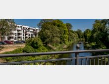 Blick von einer Brücke auf einen Fluss mit Booten, umgeben von grünen Bäumen. Links befinden sich moderne Wohngebäude mit Balkonen und einem Parkplatz davor.