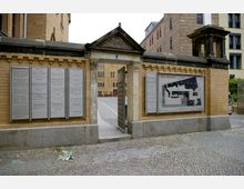 Eingang zur Kulturbrauerei in Berlin mit einer gelben Backsteinmauer und einer geöffneten Toranlage. An der Mauer sind Hinweisschilder mit Informationen zu den Einrichtungen des Geländes sowie ein Lageplan angebracht.