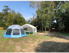 Ein offenes Gelände mit zwei Zelten, einem blauen Kuppelzelt und einem weißen Pavillon, umgeben von hohen Bäumen und etwas Rasenfläche. Im Hintergrund ist ein Holzzaun und dichter Baumbestand zu sehen.