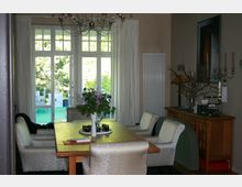 Esszimmer mit einem Holztisch und gepolsterten Stühlen, dekoriert mit einer Vase voller Blumen in der Mitte. Im Hintergrund sind bodentiefe Fenster mit Blick auf eine Terrasse und einen Garten zu sehen.