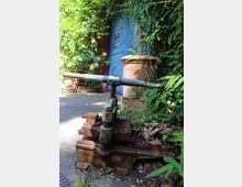 Verwachsener Gartenweg neben einer blau gestrichenen Wand mit Schild, umgeben von dichtem Grün und gelben Rosen. Im Vordergrund steht ein rostiges Metallgerät mit Hebel auf dem Boden neben einem großen Terrakottatopf.