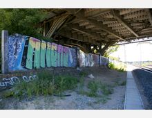 Ansicht eines Bereichs unter einer Brücke mit sichtbaren Metallträgern. Eine Mauer entlang des Bahndamms ist mit großflächigen, bunten Graffiti bemalt, während im Vordergrund überwuchertes Gelände und ein Gehweg sichtbar sind.
