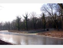 Gefrorener Fluss in einer winterlichen Parklandschaft mit kahlen, teils von Efeu bewachsenen Bäumen. Im Hintergrund verläuft eine kleine Brücke über den Fluss.