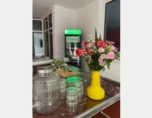Ein moderner Innenraum mit einem gedeckten Tisch im Vordergrund, der eine gelbe Vase mit frischen Blumen, ein großes Glasgefäß und mehrere gestapelte Gläser zeigt. Im Hintergrund steht ein beleuchteter Kühlschrank mit durchsichtiger Front, umgeben von großen Fenstern.