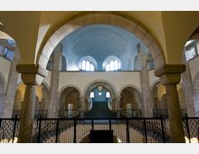 Innenraum einer historischen Kirche mit einer Mischung aus Rundbögen und Ornamenten. Sichtbar sind ein blaues, mit Mustern verziertes Gewölbe, eine zentrale Uhr und steinerne Säulen mit gedrehten und kantigen Details.