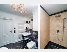 Modernes Badezimmer mit dunklem Marmorboden, einem weißen Waschbecken auf Metallgestell, einer bodengleichen Dusche mit beigefarbenen Natursteinwänden, einem Heizkörper und einem Spiegel mit zwei schwarzen Wandlampen. Die Deckenleuchte ist verziert mit einem chromfarbenen Design.