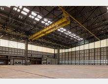 Große Industriehalle mit hohen Decken, Stahlträgern und einem gelben Deckenkran. Die Wände bestehen aus lichtdurchlässigen Paneelen und großen Toren, der Boden ist eine ebene Betonfläche.