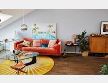 Wohnzimmer in modernem Stil mit hellblauer Wandfarbe, einem orangefarbenen Sofa mit bunten Kissen, einem runden Glastisch auf einem gelben Teppich und einem großen gerahmten Bild mit Pool-Motiv. Im Raum stehen außerdem eine Pflanze in einem Korb, ein Holzmöbelstück, ein kleiner schwarzer Beistelltisch mit Deko sowie eine silberne Hängelampe.