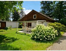 Ein Einfamilienhaus mit Holzverkleidung und Satteldach, umgeben von einem gepflegten Garten. Im Vordergrund ist eine Terrasse mit Gartenmöbeln und ein großer Strauch mit weißen Blüten zu sehen, links befindet sich ein kleiner Nebenschuppen.