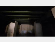 Ein dunkler Kellerraum mit sichtbaren grünen Holzbalken an der Decke, weißen verputzten Ziegelwänden und einer Teilung durch gemauerte Pfeiler. Die Beleuchtung ist schwach, wodurch der Raum düster wirkt.