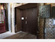 Eine industrielle Eingangsszene mit einer braunen Holztür in einem Ziegelsteinraum. Links befindet sich ein rot-grünes Türfenster, rechts eine alte Schalttafel mit sichtbaren Schaltern und Pfeildiagrammen, sowie freiliegende Rohre an der Wand.