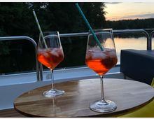 Zwei Gläser mit orangefarbenem Getränk und Strohhalmen stehen auf einem runden Holztisch auf einem Boot, im Hintergrund ein ruhiger See mit Bäumen und ein Himmel bei Sonnenuntergang.
