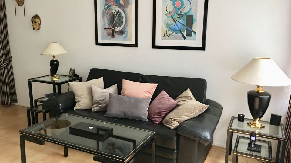 Wohnzimmer mit schwarzem Ledersofa und vielen Kissen, davor ein Glascouchtisch auf hellem Holzfußboden. An der weißen Wand hängen zwei gerahmte abstrakte Bilder; rechts und links stehen Beistelltische mit Lampen, daneben sind dekorative Masken an der Wand angebracht.
