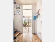 Flur eines modernen Hauses mit Holzboden, der in einen hellen Wohnraum mit großen Fenstern und Blick auf einen Garten führt. An den Wänden des Flurs befinden sich Bilderrahmen und ein Wandorganizer, eine Stehlampe steht rechts neben der Tür.