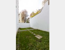 Kleiner Innenhof mit einer Rasenfläche, durchzogen von einem schmalen Weg aus rechteckigen Steinplatten. Die Umrahmung besteht aus hohen weißen Wänden, und im Hintergrund sind Bäume sowie Mehrfamilienhäuser sichtbar.