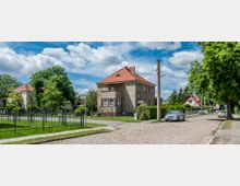 Wohnviertel mit Kopfsteinpflasterstraße, umgeben von Einfamilienhäusern mit roten Ziegeldächern und gepflegten Gärten. Links im Bild steht ein zweistöckiges Wohnhaus mit Zauneinfriedung, und rechts säumen Bäume die Straße, wo mehrere parkende Autos sichtbar sind.