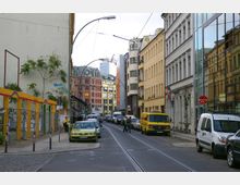 Straßenszene in einem städtischen Viertel mit mehrstöckigen Gebäuden aus verschiedenen Architekturstilen, teilweise mit Graffiti bemalt. Die Straße ist von parkenden Autos gesäumt, Gleise verlaufen in der Mitte, und Passanten bewegen sich zwischen den Häusern.