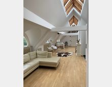 Offener Dachgeschossraum mit heller Holzbodenfläche, einem beigefarbenen Ledersofa, einem Schreibtischbereich, zwei weißen Stühlen, einem Fellteppich und mehreren großen Dachfenstern mit Holzverkleidung. Die schrägen Wände und sichtbaren Balkenelemente verleihen dem Raum eine moderne, luftige Atmosphäre.