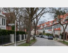 Gepflegte Wohnstraße mit Reihenhäusern im traditionellen Stil, erkennbar an roten Ziegeldächern und weißen Fassaden mit grünen Fensterläden. Die Straße ist gepflastert, gesäumt von Bäumen, geparkten Autos und gepflegten Vorgärten mit Zäunen. Ein Straßenschild weist auf die "Wachtelstraße" hin.