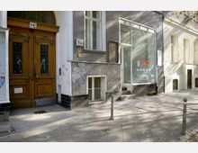 Wohnhausfassade in einer städtischen Straße mit einer großen Holzeingangstür und verzierten Glaseinsätzen links sowie einem Schaufenster mit Glasfront rechts. Der Gehweg ist mit Steinen gepflastert, teilweise durch Poller und Bäume begrenzt, und weist Schatten von umliegenden Bäumen auf.