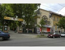 Ein Eingang zu einem Biergarten mit der Beschilderung "Prater Garten", flankiert von Bäumen und Backsteinpfeilern. Rechts daneben ein Gebäude mit gelber Fassade, Graffiti und einer dekorativen Metallrad-Installation, daneben parkende Autos und eine Straßenbahnlinie im Vordergrund.