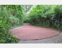 Ein kleiner, rundlicher Platz mit rötlichem Bodenbelag, umgeben von dichtem Grün aus Büschen und Bäumen in einem parkähnlichen Gelände. Im Vordergrund sind Pflastersteine sichtbar, die in den Platz übergehen.