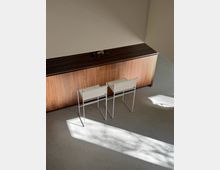 Minimalist interior corner with a long wood-paneled cabinet against a plain wall and two small metal-framed side tables in front. Sunlight streams across a light gray floor, casting sharp rectangular shadows.