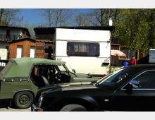 Wohnwagensiedlung mit geparkten Fahrzeugen im Vordergrund, darunter ein grünes Cabrio und ein dunkles Auto. Im Hintergrund sind ein weißer Wohnwagen, Holzhütten und Bäume sichtbar; daneben steht eine Person mit einem blauen Eimer und rotem Pullover.