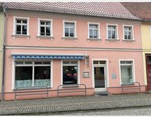 Zweistöckiges, rosafarbenes Stadthaus mit roten Ziegeln, weißen Fensterrahmen und mehreren Sprossenfenstern; im Erdgeschoss befindet sich ein kleines Café mit blau-weiß gestreifter Markise und großen Schaufenstern. Vor dem Gebäude verläuft ein gepflasterter Gehweg mit niedrigen Metallbügeln entlang der Straße.
