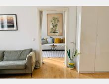 Wohnzimmer mit Holzboden, einem grauen Sofa links und einem weißen Schrank rechts. Durch eine Türöffnung mit Vorhängen ist ein weiterer Raum sichtbar, der mit einer weißen Sitzbank, bunten Kissen, einem kleinen runden Tisch und einem Palmenbild an der Wand dekoriert ist.