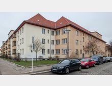 Wohnblock in einer Straßenecke mit hellem Putz und roten Ziegeldächern, umgeben von spärlich begrünten Bäumen und parkenden Autos entlang einer gepflasterten Straße. Im Vordergrund stehen mehrere Straßenschilder und eine Laterne.