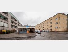 Straßenszene mit mehrstöckigen Wohngebäuden, einem Zugang zur U-Bahn mit dem Schild „Gleiwitzer Straße“ und abgestellten Fahrzeugen am Straßenrand. Der linke Gebäudekomplex ist grau mit Balkonen, der rechte gelb mit schlichten Fassaden, die Umgebung wirkt urban.