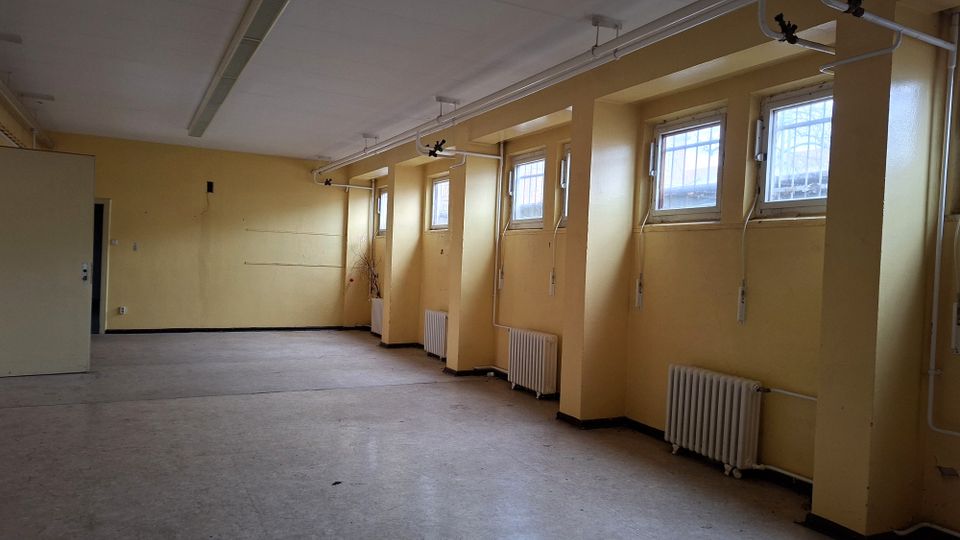 Leerer, länglicher Innenraum mit gelb gestrichenen Wänden, mehreren hohen Fenstern in tiefen Nischen und Heizkörpern darunter. An der Decke verlaufen Rohrleitungen und lange Leuchtstofflampen über einem grauen Boden.