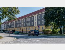 Mehrstöckiges Wohnhaus mit grauer und gelber Fassade, roten Ziegeldächern und vielen symmetrischen Fenstern entlang einer gepflasterten Straße. Vor dem Gebäude parken mehrere Autos, und Bäume säumen die Straße.