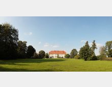 Ein weißes Herrenhaus mit rotem Ziegeldach steht am Ende einer weitläufigen, gepflegten Rasenfläche, umgeben von hohen, dicht bewachsenen Bäumen unter klarem, blauem Himmel.