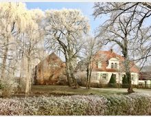 Ein großes Landhaus mit rotem Ziegeldach und weißen Fensterrahmen, umgeben von frostbedeckten Bäumen und Sträuchern; im Vordergrund ein kleiner Graben und eine Hecke. Der Himmel ist klar, und die Umgebung wirkt winterlich.