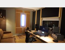 Ein moderner Tonstudiobereich mit einem großen Schreibtisch, auf dem ein Mischpult, Lautsprecher und ein Monitor stehen. Im Raum befinden sich schallabsorbierende Wandverkleidungen, eine braune Couch und ein Sichtfenster, das in einen angrenzenden Raum führt.