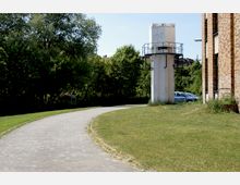 Gepflasterter Weg, der an einer Rasenfläche und einem großen weißen Mast mit Plattform vorbeiführt, rechts ein Gebäude mit Backsteinfassade; im Hintergrund Bäume und parkende Autos.