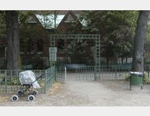 Ein kleiner Parkeingang mit grünem Metallzaun und Tor, dahinter eine Bank und Bäume vor einer roten Backsteinfassade. Im Vordergrund stehen ein Kinderwagen mit Plastikabdeckung und ein Abfalleimer.