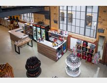 Innenraum eines Verkaufsraums in einem Industriestil-Gebäude mit großen Fenstern, Backsteinwänden und Metallelementen. Im Vordergrund stehen Flaschendisplays, daneben ein Tresen mit Spirituosen und Merchandising-Produkten.