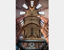 Innenansicht einer neugotischen Kirche mit rotem Ziegelmauerwerk, spitz zulaufenden Balkendecken und großen, hochgelegenen Fenstern. Im Vordergrund ist eine Empore aus Metall zu sehen, darunter eine Orgel, die von gestuften Verzierungen und Bögen umrahmt wird.