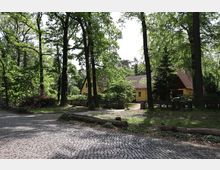 Ein gelbes Haus mit rotem Ziegeldach steht in einer ländlichen Umgebung, umgeben von hohen Bäumen und einem kleinen Zaun. Im Vordergrund ist eine gepflasterte Straße zu sehen, die von Gras und vereinzeltem Gehölz gesäumt ist.