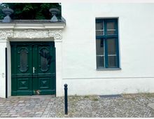 Weiße Hausfassade mit einer dunkelgrünen, verzierten Holztür links, über der die Inschrift „Gott segne das Handwerk“ in ein Relief eingearbeitet ist. Rechts daneben befindet sich ein einfaches rechteckiges Fenster mit grünen Rahmen; der Boden vor der Fassade ist mit Kopfsteinpflaster bedeckt.