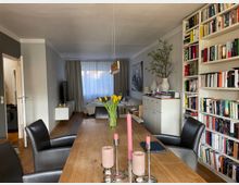 Wohnzimmer mit Essbereich in einer modernen Wohnung. Ein langer Holztisch mit schwarzen Lederstühlen steht im Vordergrund, dahinter ein Wohnbereich mit Couch, Bücherregal, Sideboard und großen Fenstern mit Vorhängen.