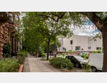 Straßenszene in einer städtischen Umgebung mit einem alten, rot-gelben Wohngebäude links, einem modernen, weißen Betongebäude rechts und einer von Bäumen gesäumten Gehwegallee. Parkende Autos, Grünflächen und Fußgänger prägen das Bild.