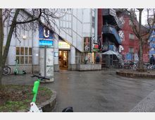 Außenansicht eines U-Bahnhof-Eingangs der Station "Prinzenstraße", mit umliegendem Gehweg, E-Scootern und Fahrrädern. Rechts daneben ein mit Graffiti bedecktes Gebäude, eine Imbissbude und Sitzmöglichkeiten unter einem Sonnenschirm.