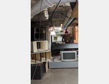 Ein Kellerraum mit sichtbaren Rohren an der Decke, einem DJ-Pult, einem alten Fernseher auf einem Podest und großen Lautsprechern an der Wand. Im Hintergrund steht eine Person mit Mütze am Mischpult, umgeben von weiterem technischen Equipment.